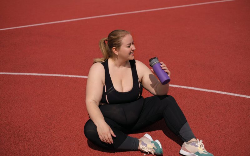 Fat, plump, obese woman, tired after exercising, drinks water from bottle while sitting on the sports red surface of ground. Happy smiling person strive to lose body weight