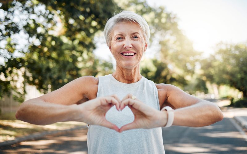 Fitness, happy and heart hands of old woman in nature after running for health, wellness and workout. Smile, motivation and peace with senior lady and sign for love, faith and training in nature.