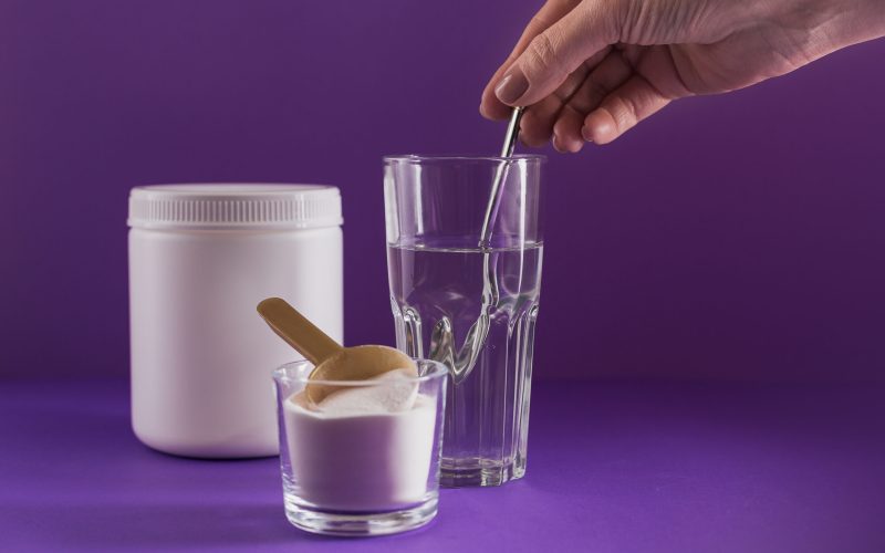 Glass with collagen dissolved in water and collagen protein powder on purple background. Woman's hand holds a spoon. Healthy lifestyle concept.