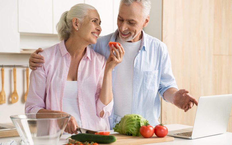 Picture of cheerful mature loving couple family standing at the kitchen using laptop computer and cooking. Looking aside.