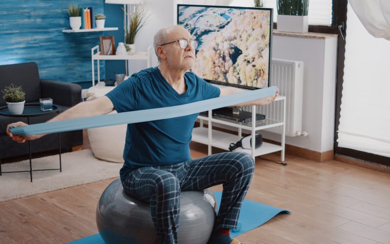 Senior man using resistance band to do physical exercise with arms while sitting on fitness toning ball. Aged person training with flexible elastic belt and workout equipment. Adult exercising